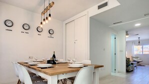 Un coin repas avec une table en bois dressée pour quatre personnes, des chaises blanches et une bouteille de champagne. On y trouve également des horloges murales indiquant le nom de différentes villes.