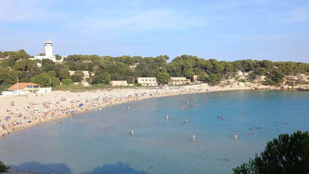 Une plage côtière avec un phare, une eau d’un bleu limpide et une côte rocheuse.