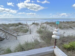 Vista de la playa con una escalera de madera que baja a la arena, una barrera de cuerda y una pila de piedras con un objeto azul encima.