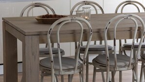 Een houten eettafel met zes stoelen, elk met een wit frame en grijze kussens. Op de tafel staat een schaal met gedroogde bloemen en een kaars.