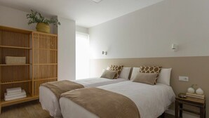 Een hotelkamer met twee bedden, een houten garderobe, een nachtkastje met een vaas en een raam met natuurlijk licht.