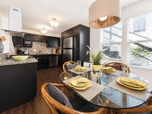 A modern kitchen with dark cabinetry, stainless steel appliances, and a dining area with a glass table and wooden chairs.