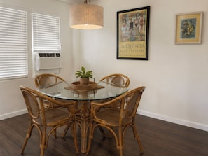 A dining area with a glass table surrounded by wicker chairs, a potted plant, and a wall-mounted air conditioner.