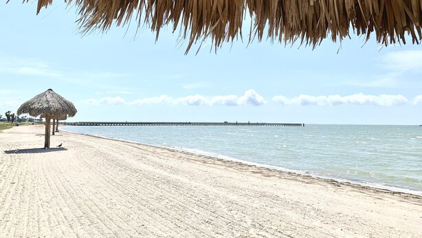 A beach with a thatched umbrella and a pier in the background. - The Sand Piper-Pier, Pool, Bay View & Boat Launch (Rockport)