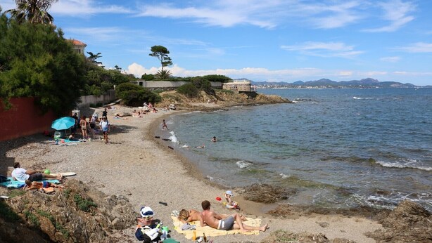 A beach with people sunbathing and swimming, a rocky shoreline, and a building with a red roof in the background. - Seaside apartment (Saint-Aygulf)