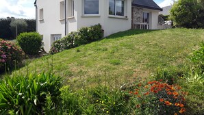 A two-story house with a sloped roof, a front porch, and a garden with various plants and flowers. - HOLIDAY HOUSE WITH STUNNING SEA VIEWS (Locquirec)