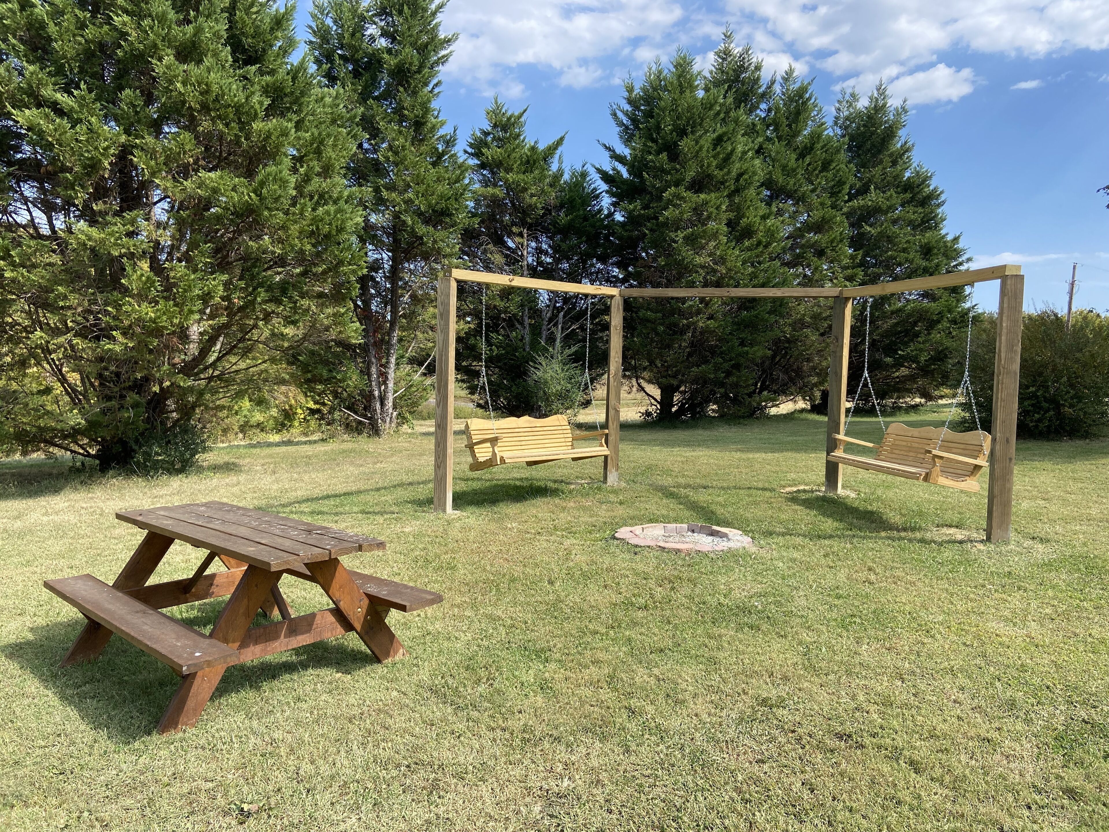 A wooden swing set, picnic table, and fire pit in a grassy park area with trees.