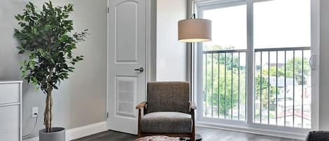 A room with a large window, a potted plant, a chair, a white dresser, and a lamp.