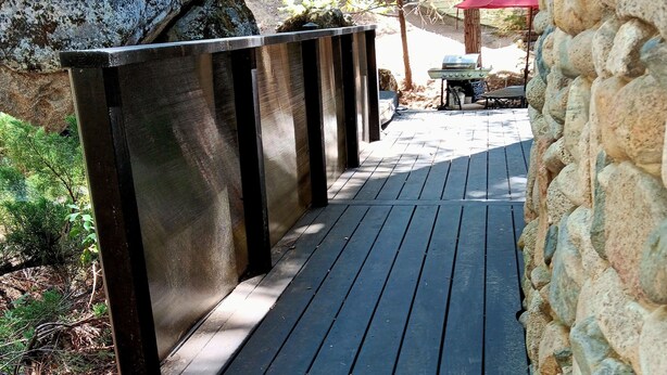 Una terraza de madera con escalones que conducen a una zona de estar, rodeada de muros de piedra y vegetación.