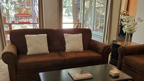 Un sofá de cuero marrón con dos almohadas blancas con textura, una mesa de centro negra con un libro y un objeto blanco, y un jarrón con flores blancas.