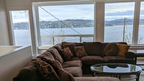 A modern living room with a large sectional sofa, a glass coffee table, and a view of a lake and hills. - Stunning views of Lake Washington (Seattle)