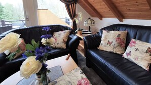 Ein gemütliches Wohnzimmer mit zwei schwarzen Ledersofas, jeweils mit floralen Kissen, einer Holzbalkendecke und Blick ins Freie durch ein großes Fenster.