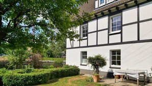 Un bâtiment à deux étages, doté d’une façade blanche et de bordures sombres. On y trouve un petit jardin avec un banc et des plantes en pot.