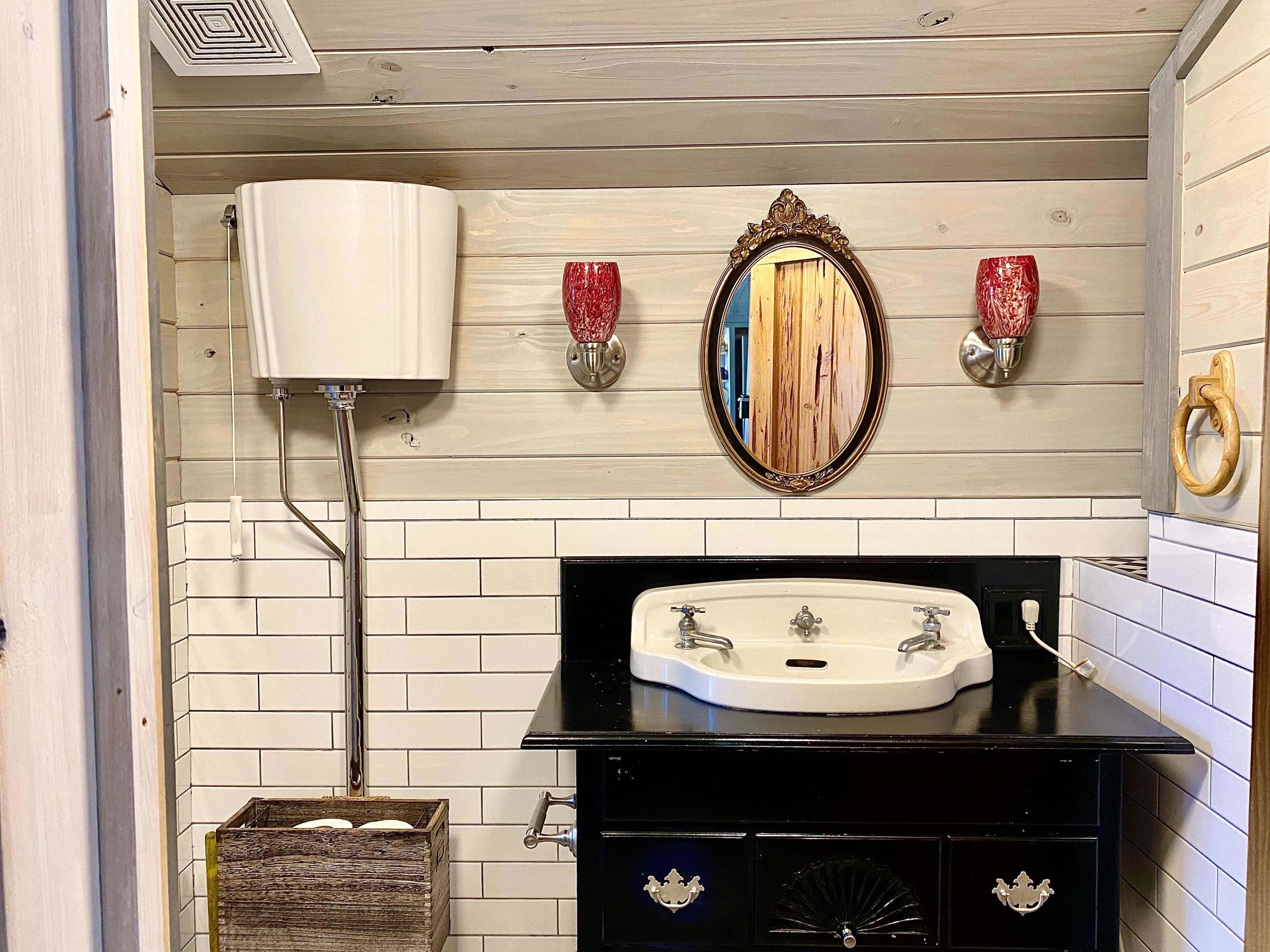 Un baño con pared de azulejos blancos, un lavamanos negro con dos grifos, un mueble de madera debajo y un espejo con marco decorativo.