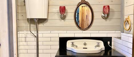 Un baño con pared de azulejos blancos, un lavamanos negro con dos grifos, un mueble de madera debajo y un espejo con marco decorativo.