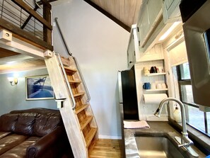A compact kitchen with a sink, a wooden ladder, and a shelf with kitchenware.