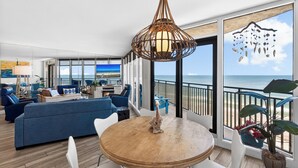 Een ruime woonkamer met een groot raam met uitzicht op zee, een ronde houten tafel en een hanglamp.