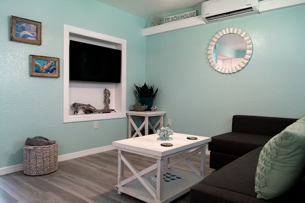 A living room with a flat-screen TV, a small table, a couch, and a wall-mounted mirror.
