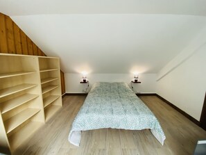 Un pequeño dormitorio en el ático con techo inclinado, una estantería de madera, una colcha estampada y dos lámparas de noche.