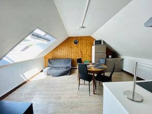 A compact, modern loft bedroom with a sloped ceiling, a built-in wooden wall, a small kitchenette, and a cozy seating area with a sofa and chairs. - Résidence les pâquerettes N4 (Mont-Dore)