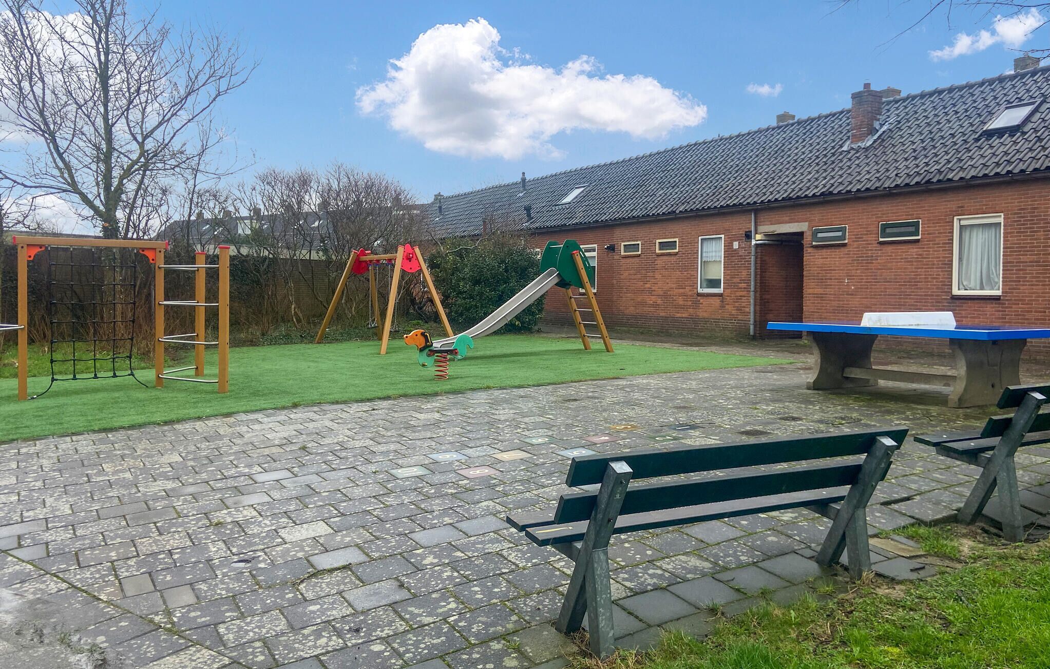 Une aire de jeux avec des balançoires et un toboggan, un bâtiment en briques doté de fenêtres et une zone pavée avec des bancs.