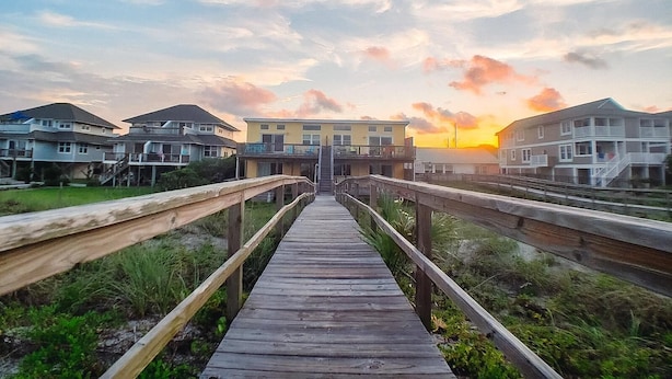Een houten looppad dat naar een rij strandhuizen leidt, met een zonsondergang op de achtergrond.