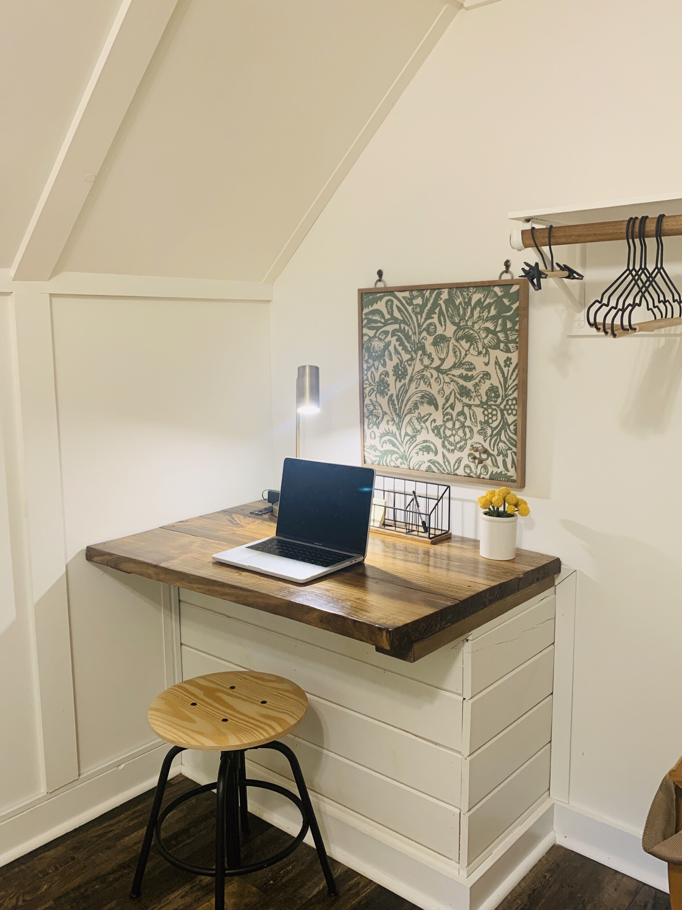 A corner desk with a laptop, a small stool, a wall-mounted shelf with a vase of flowers, and a hanging coat rack.