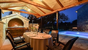 An outdoor dining area with a stone fireplace, wooden chairs, and a pool in the background.