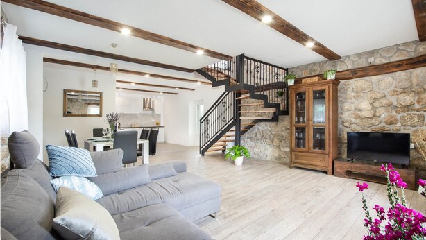 A spacious living room with a grey sofa, a stone fireplace, a wooden staircase, and a kitchen in the background.