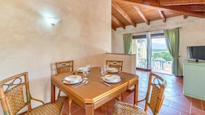 Uma sala de jantar com mesa de madeira e cadeiras de vime, uma televisão sobre um móvel e vista para o exterior através de uma porta de vidro.