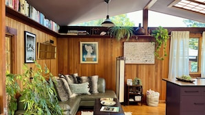 A cozy living room with a sectional sofa, a coffee table, and a bookshelf filled with books. There are several plants, a framed picture on the wall, and a window with curtains. - The Bathhouse: Boho Modern + Hot Tub + Water View (Mill Valley)