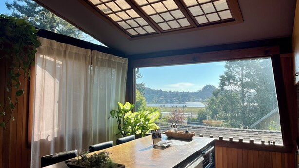 A room with a wooden table, chairs, a window with a view of a lake and mountains, and a ceiling with a grid pattern. - The Bathhouse: Boho Modern + Hot Tub + Water View (Mill Valley)