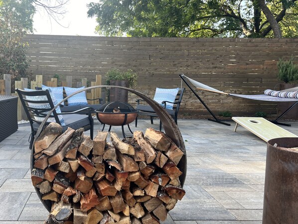 Eine Terrasse im Freien mit Feuerstelle, gestapeltem Brennholz, Liegestühlen und einer Hängematte.