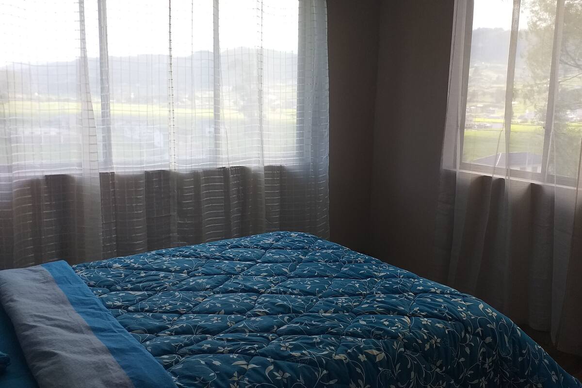 Ett sovrum med en säng, ett fönster med tunna, genomskinliga gardiner och utsikt över ett grönskande landskap.