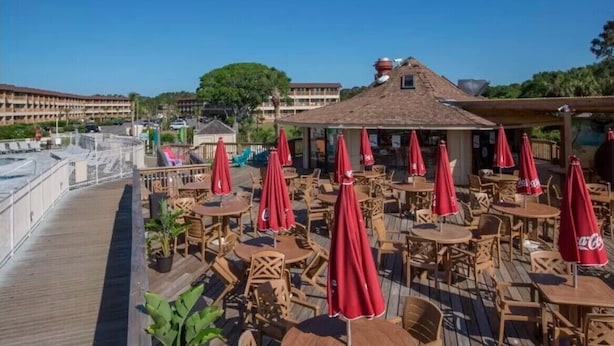 An outdoor patio with wooden tables and red umbrellas, overlooking a waterfront area.