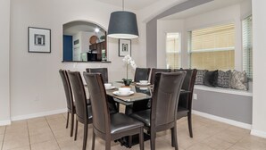 Un coin repas avec une table dressée pour six personnes, entourée de chaises de couleur brun foncé. Un grand lustre est suspendu au-dessus de la table, et une fenêtre avec des stores se trouve à droite.
