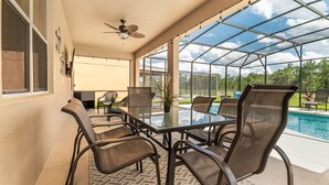 Un patio couvert avec une table et des chaises en verre, un ventilateur de plafond et une vue sur une piscine et la verdure environnante.