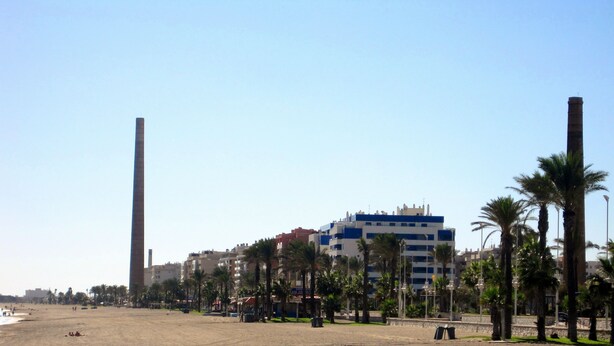 A beach with buildings, palm trees, and a tall chimney. - Paseo Marítimo Antonio Banderas (Málaga)