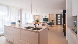 Een moderne keuken met een centraal eiland, een eethoek met tafel en stoelen, een woonkamer met bank en televisie, en een keukenblad met apparatuur.