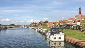 A canal with boats docked, a row of houses with red roofs, and a tall chimney on a building.