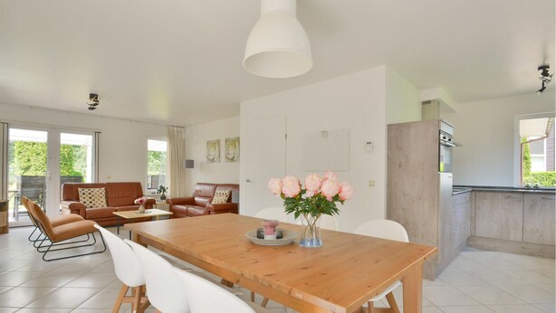 A spacious living area with a wooden dining table, white chairs, a sofa, and a kitchen in the background.