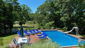 Une piscine rectangulaire avec un toboggan, entourée de chaises longues bleues et d’une table avec des chaises.