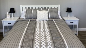 A neatly made bed with a patterned bedspread and pillows, flanked by two bedside tables with lamps and a small drawer. A landscape painting hangs above the bed. - Home on the Range (Hurricane)