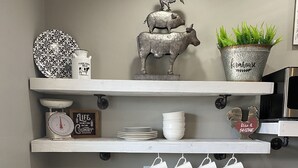 A kitchen with white cabinetry, a stainless steel sink, a stove, and a dishwasher. There are decorative items on the shelves, including a rooster, a pig, and a plant. - Home on the Range (Hurricane)