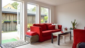 Een lichte woonkamer met een rode bank, een rieten stoel, een klein tafeltje en uitzicht op een houten schutting en groen.