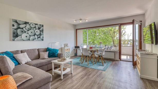 Ein modernes Wohnzimmer mit grauem Sofa, einem kleinen weißen Tisch und einem Essbereich mit Tisch und Stühlen. Ein großes Fenster bietet Blick auf Bäume und es gibt einen Balkon.