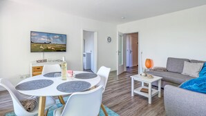 Ein modernes Wohnzimmer mit einem runden Tisch, weißen Stühlen, einem grauen Sofa und einem an der Wand befestigten Fernseher.