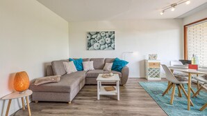 Ein modernes Wohnzimmer mit einem grauen Ecksofa, einem kleinen weißen Beistelltisch, einem blauen Teppich und einem Essbereich mit weißem Tisch und Stühlen.