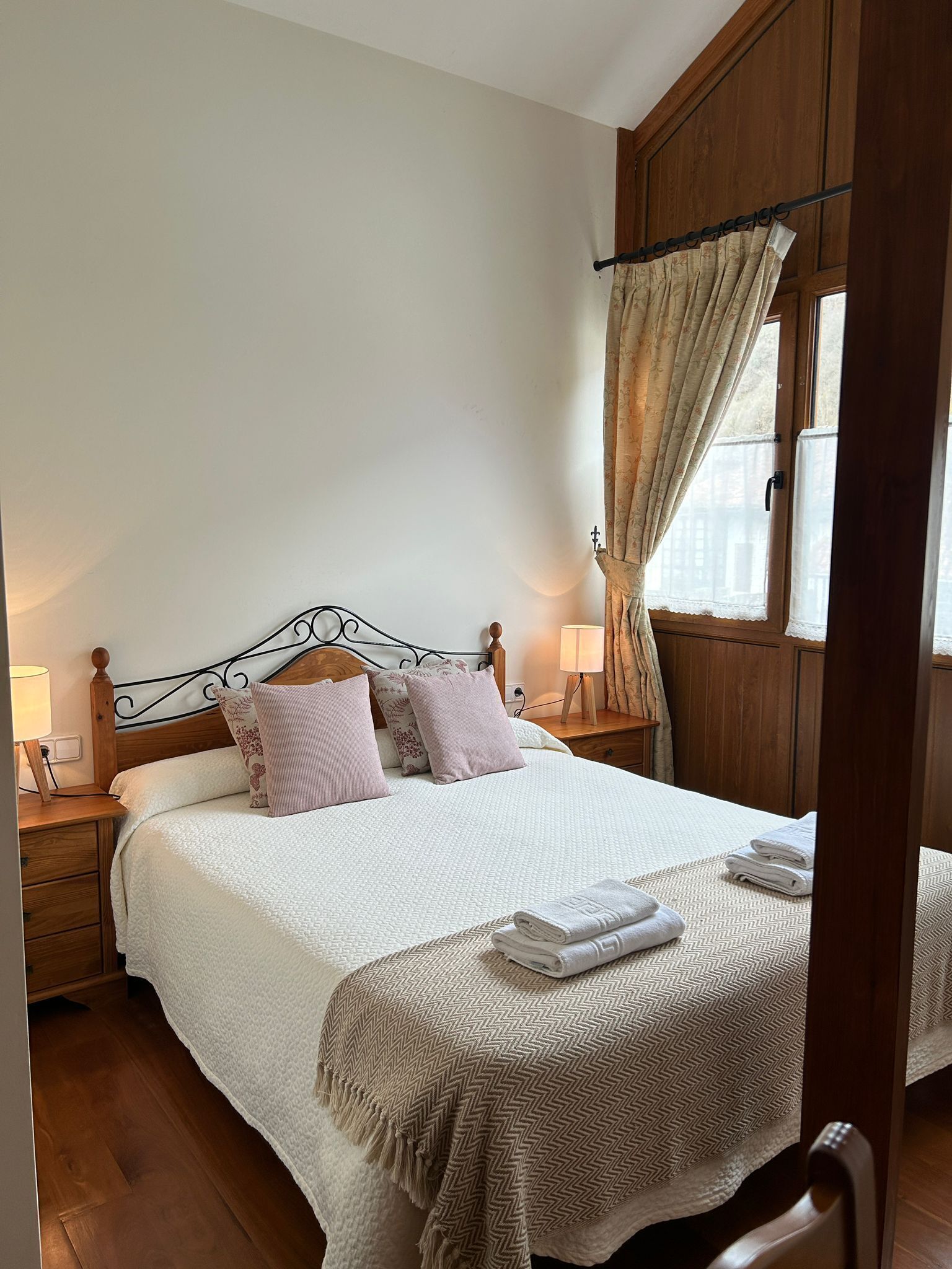 A neatly made double bed with a white and beige patterned bedspread, two pillows, and a folded towel on the side. There is a wooden nightstand with a lamp and a wooden chair. The room has wooden walls and a window with curtains.