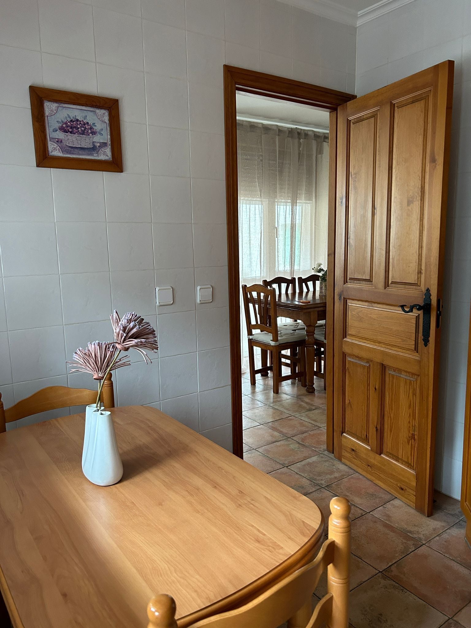 Un comedor con una mesa de madera, sillas y un jarrón con flores. Una puerta de madera da a otra habitación.